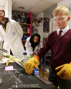 Kent scientists in lab coats watch a school child [ick up a frozen train with large yellow gloves on